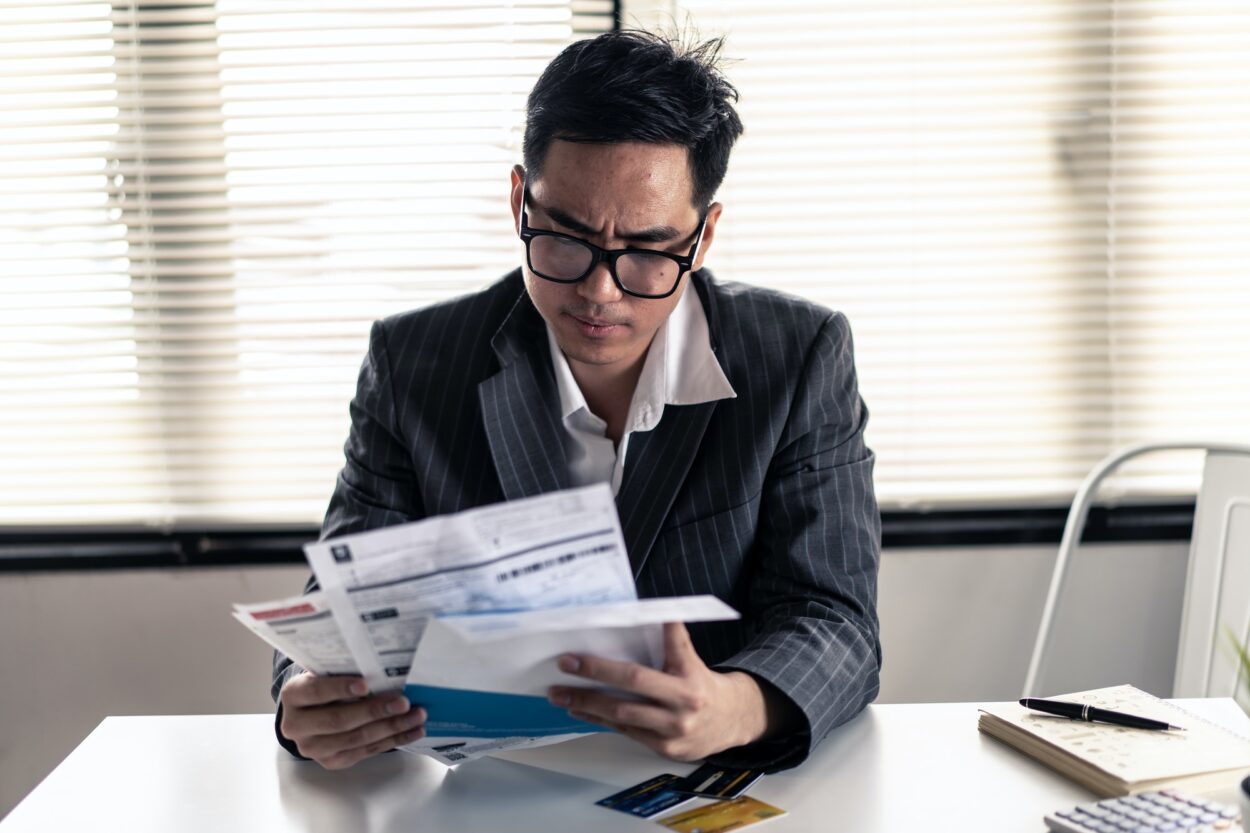 Stressed young Asian man with business office wear holding bills and another hand on head,