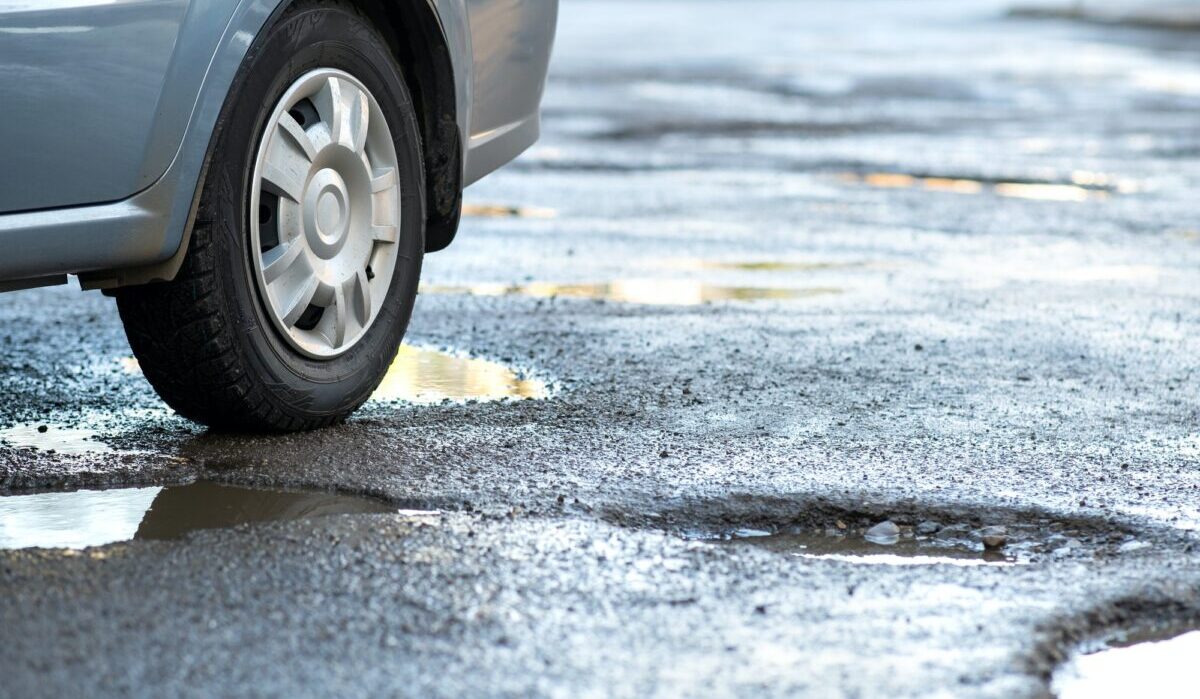 Gros plan sur une roue de voiture sur une route en très mauvais état avec de gros nids de poule remplis d'eau de pluie sale