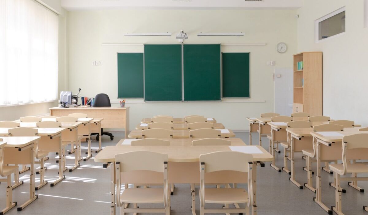 aula escolar vacía con junta escolar verde y pupitres con sillas.