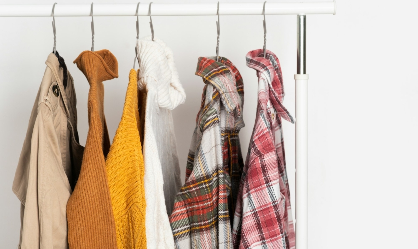 Herbstliche Vintage-Kleidung hängt auf Kleiderbügeln am Regal. Beigefarbene Trenchcoats, Pullover, karierte Hemden