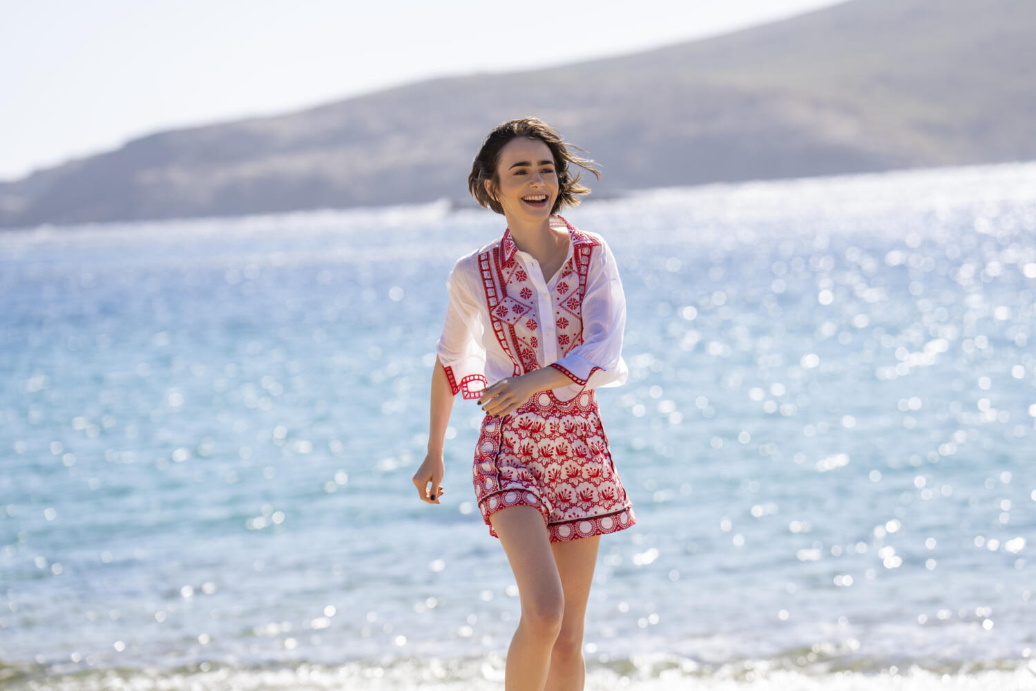Emily in Paris 6, Emily corre sorridendo su una spiaggia davanti al mare e alle montagne